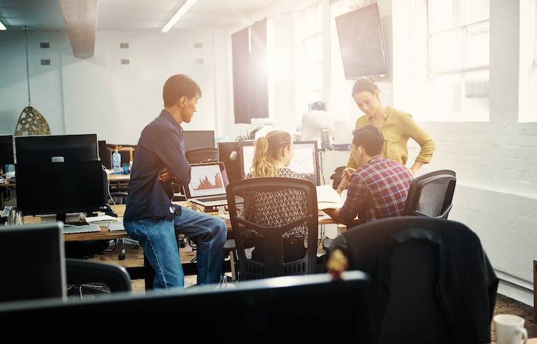 A team of professionals in a sunlit room surround a desk with a computer and discuss a successful marketing campaign