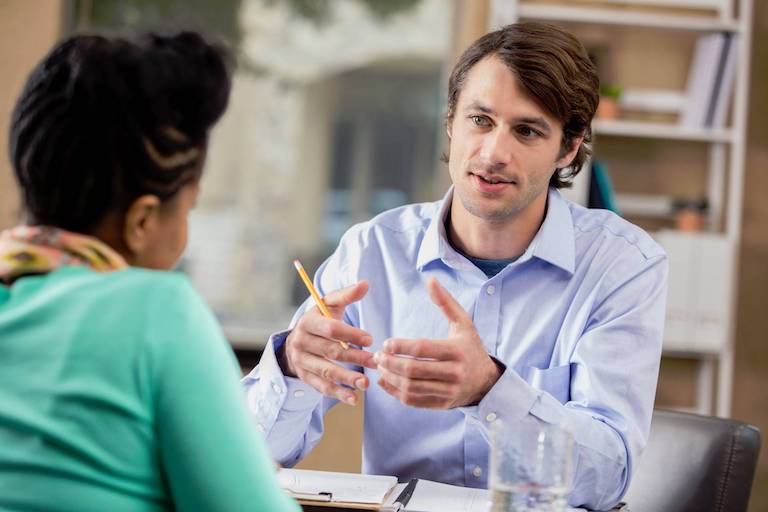 A professional man and woman sit across from each other as the man gestures meaningfully about a business topic