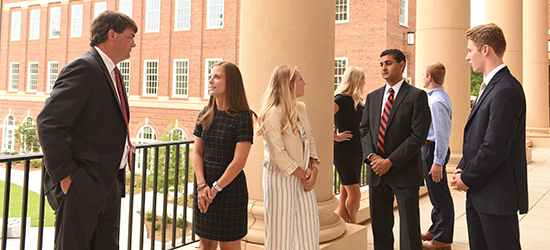 A gathering of formally dressed business students stand on the veranda of Amos Hall with a professional mentor 