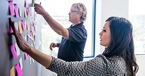 Professional woman in the foreground with a professional man reaching out to a wall with post-it notes 