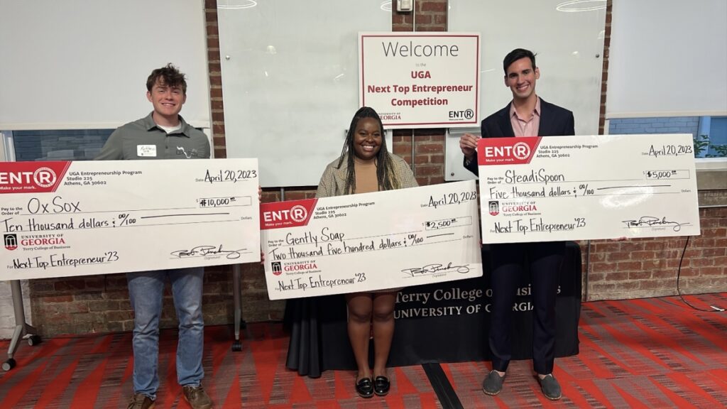 Three students stand with large checks after placing in the Next Top Entrepreneur competition. 