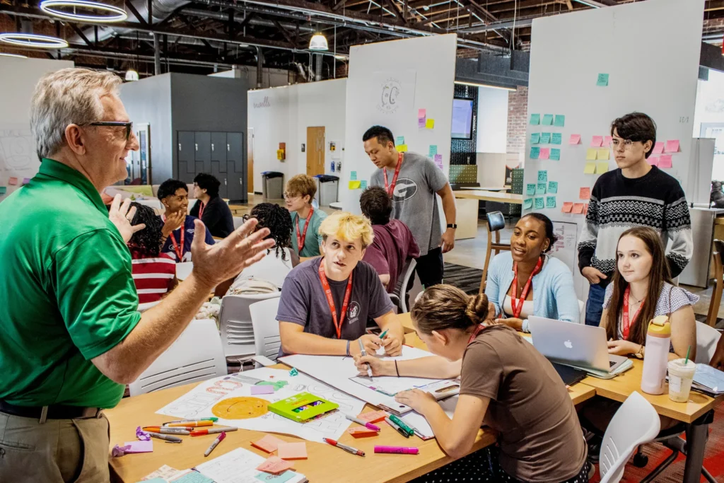 a professor talks to a group of young college students during a workshop. they are sitting at large tables and are surrounded by dry erase boards covered with notes and Post-its