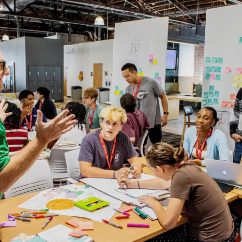 a professor talks to a group of young college students during a workshop. they are sitting at large tables and are surrounded by dry erase boards covered with notes and Post-its