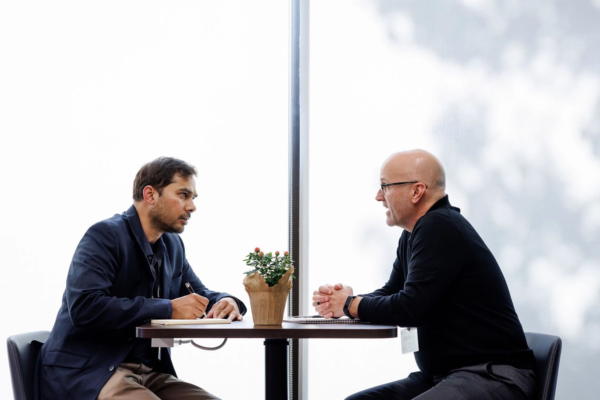 two men conversing at a small table by a window