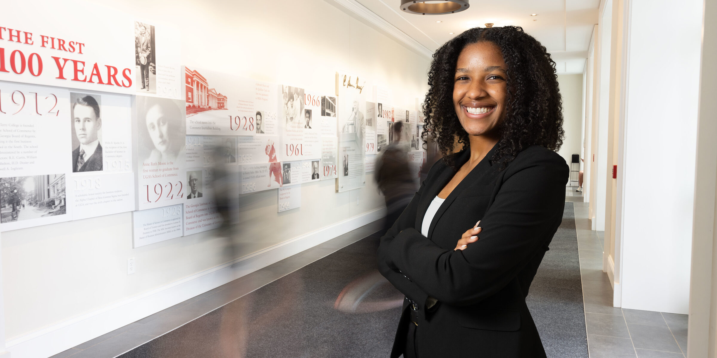 Kendall Valdry stands in a hallway in front of a Terry College of Business centennial timeline wall display at the UGA Business Learning Community