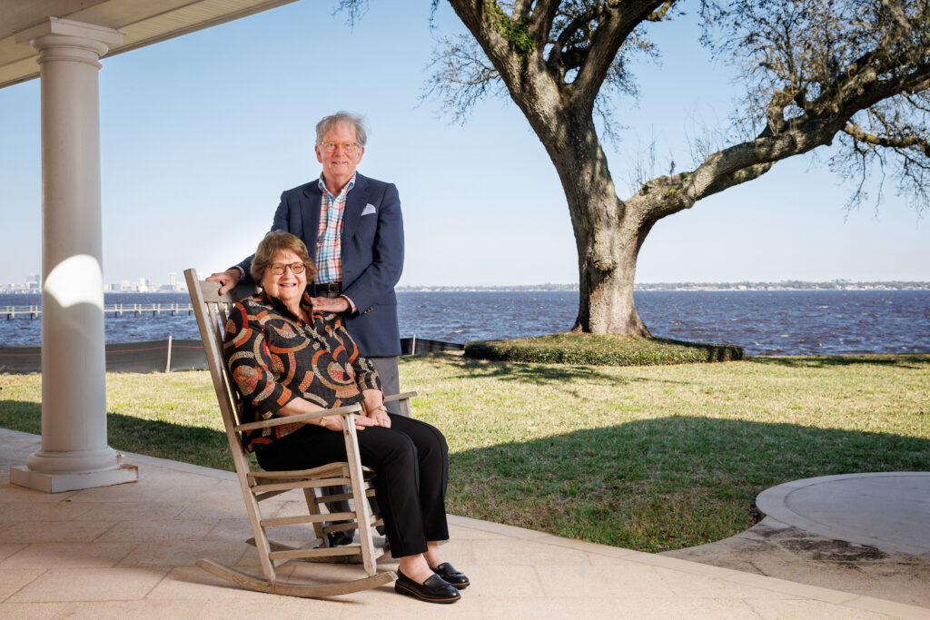 man stands next to wife seated in rocking chair on patio near waterfront