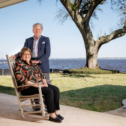 man stands next to wife seated in rocking chair on patio near waterfront