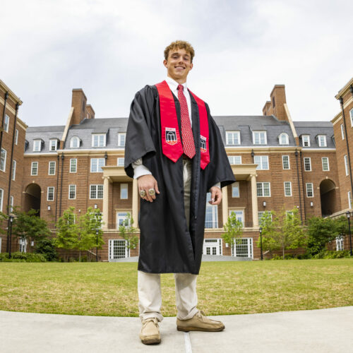 male student dressed in graduation regalia with brick buildings in background