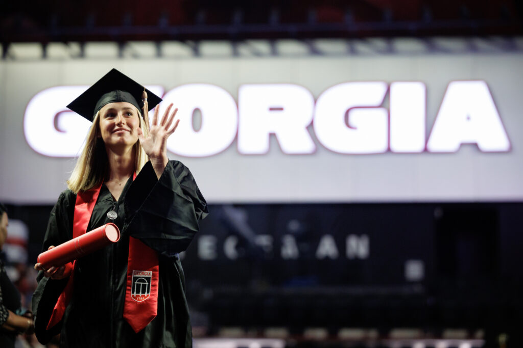 female graduate waves as she walks across stage with illuminated "GEORGIA" letters behind her