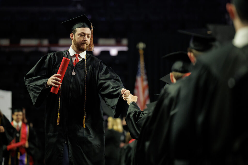 male graduate fist-bumps another graduate as he returns to his seat