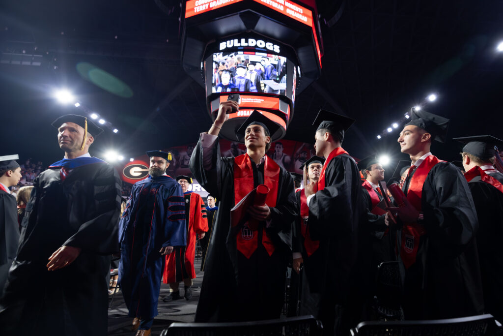 2026.05.08_rh_Undergrad_Convocation_003 One graduate pauses for a selfie while a crowd of graduates exit the floor of Stegeman Coliseum after Terry College Undergraduate Convocation on May 8.