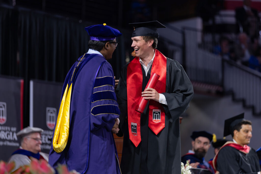 2026.05.08_rh_Undergrad_Convocation_080 Dean Santanu Chatterjee, in full academic regalia, shakes a graduate's hand while handing him a diploma tube on stage at Stegeman Coliseum during Terry College Undergraduate Convocation on May 8.
