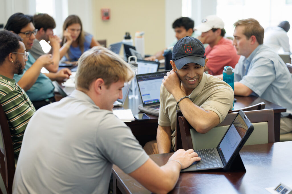 A group of students collaboratively works on laptops in a classroom setting, with one individual smiling while leaning on the table.