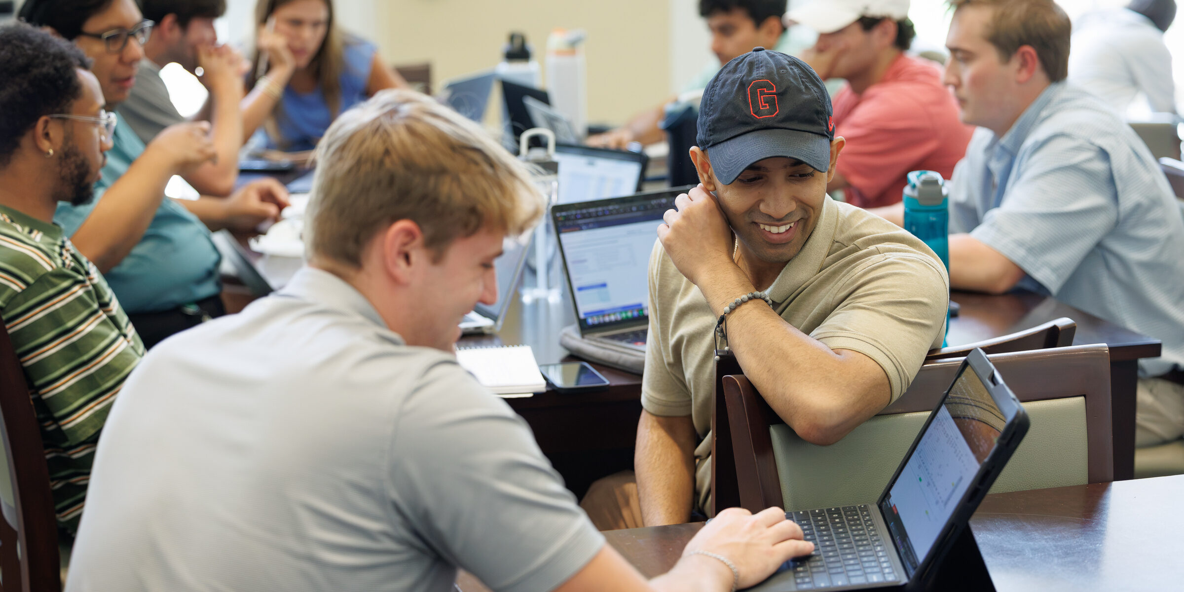 A group of students collaboratively works on laptops in a classroom setting, with one individual smiling while leaning on the table.