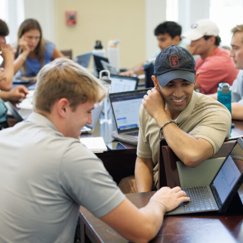 A group of students collaboratively works on laptops in a classroom setting, with one individual smiling while leaning on the table.