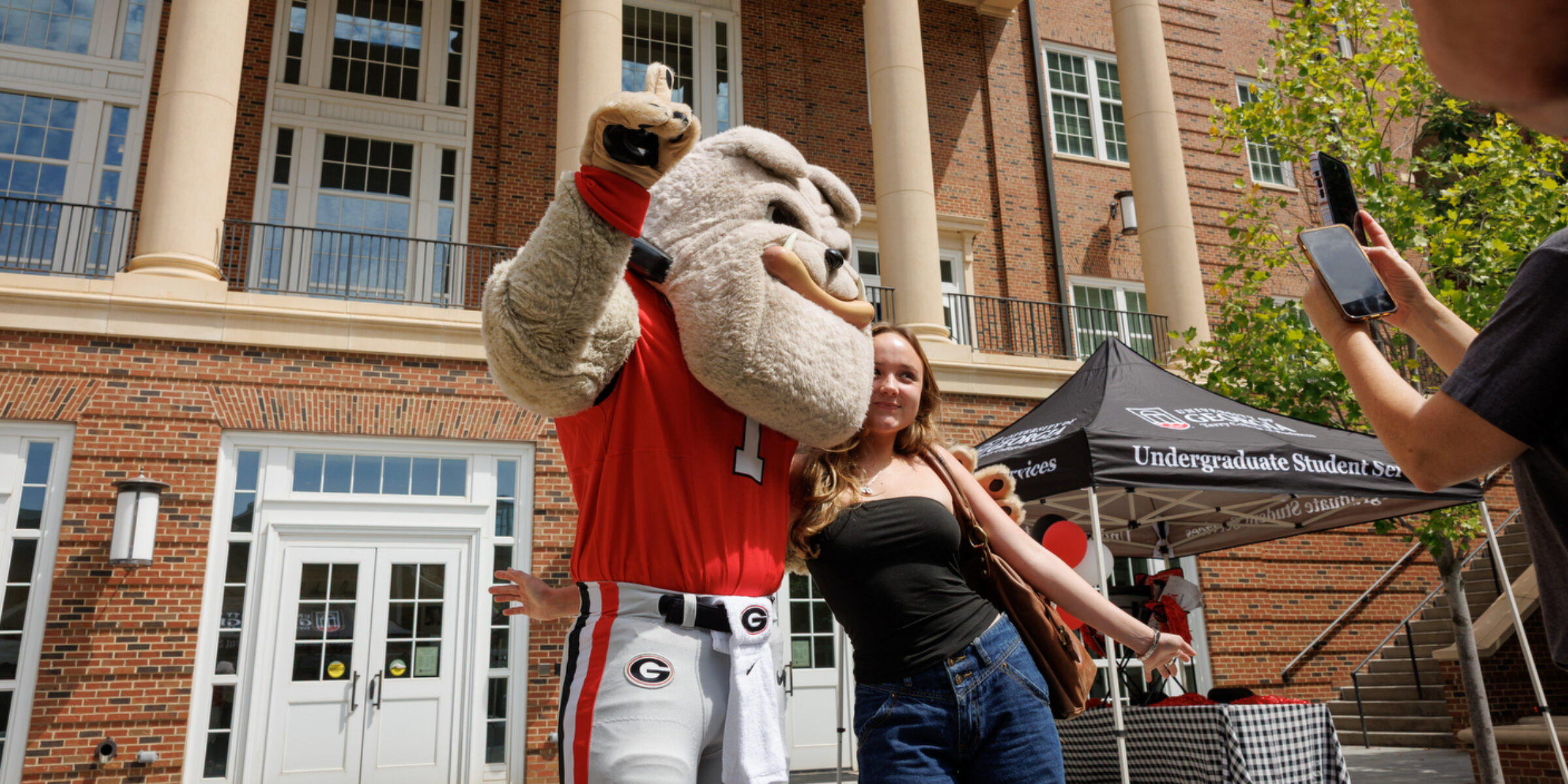 Hairy Dog gives a student a side hug underneath a Back to Business Banner hanging from Amos Hall in the Business Learning Community.