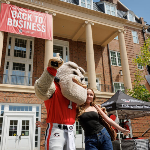 Hairy Dog gives a student a side hug underneath a Back to Business Banner hanging from Amos Hall in the Business Learning Community.