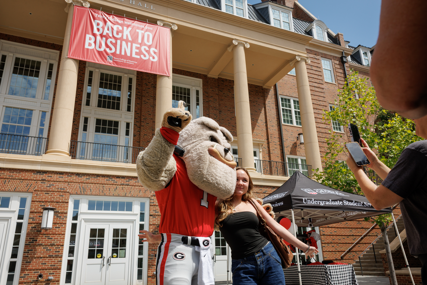 Hairy Dog gives a student a side hug underneath a Back to Business Banner hanging from Amos Hall in the Business Learning Community.