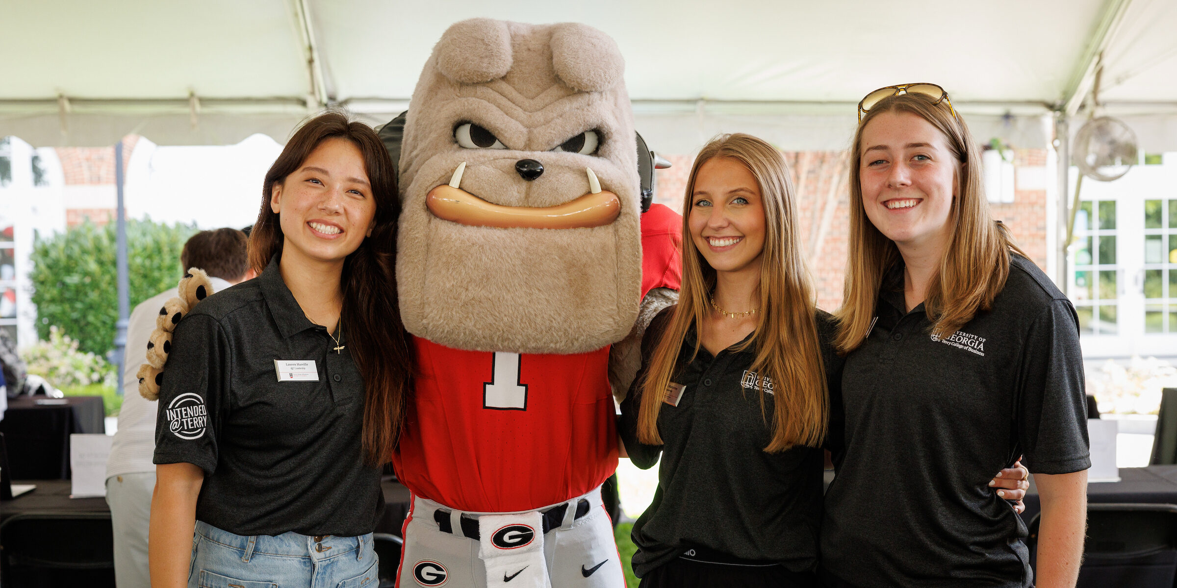 Three individuals pose with UGA Mascot Harry Dawg wearing a red jersey and white pants under a tent.
