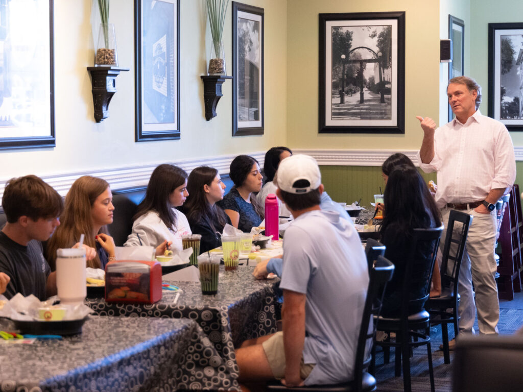 Scott Deviniey stands and speaks to a group seated at tables in a restaurant, with framed artwork and decorative plants on the walls. Chicken Salad Chick CEO teaching at Terry College
