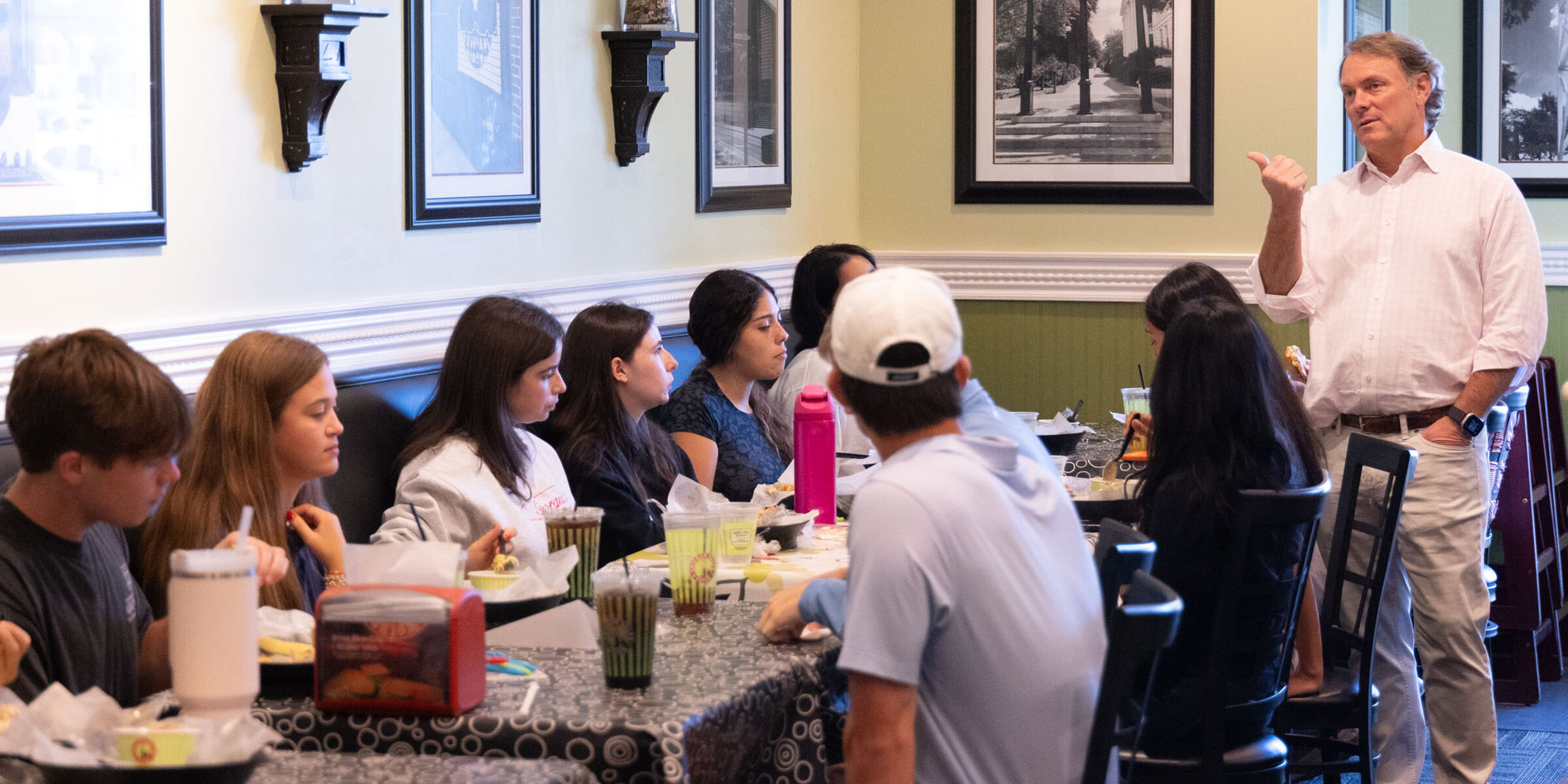 Scott Deviniey stands and speaks to a group seated at tables in a restaurant, with framed artwork and decorative plants on the walls. Chicken Salad Chick CEO teaching at Terry College
