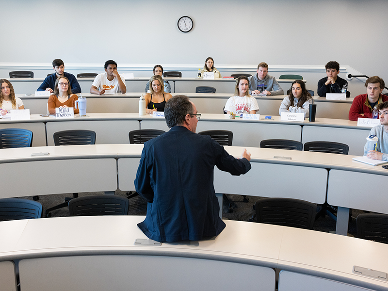 Neil Bendle sits on a desk at the front of a classroom teaching students. The photo is taken from the back so his face is not visible.