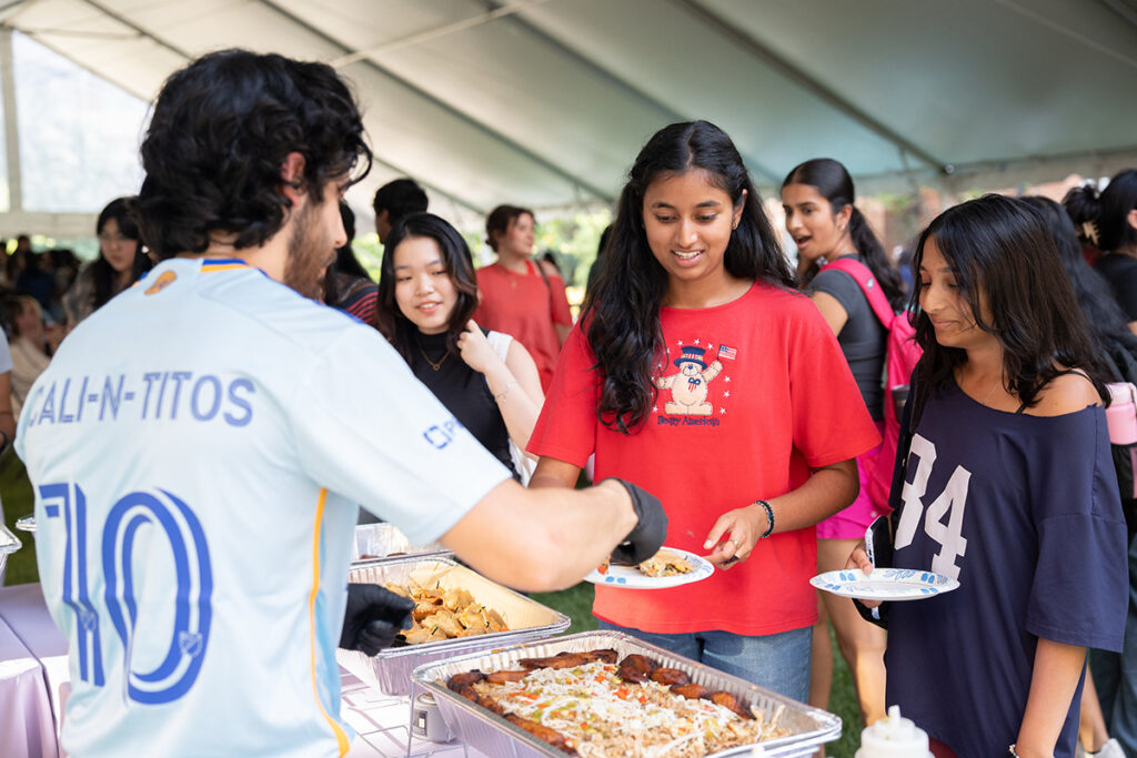 BlockParty_2026_10 A person in a sports jersey serves food from a tray to a young individual wearing a red shirt at an outdoor event under a tent.
