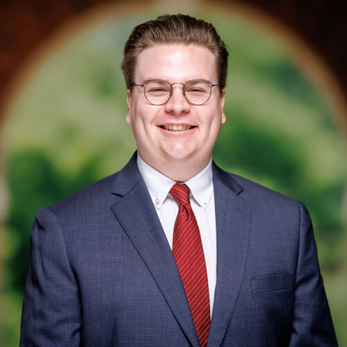 A person wearing glasses, a suit, and a red tie smiles at the camera against a blurred background featuring an arched doorway with greenery.