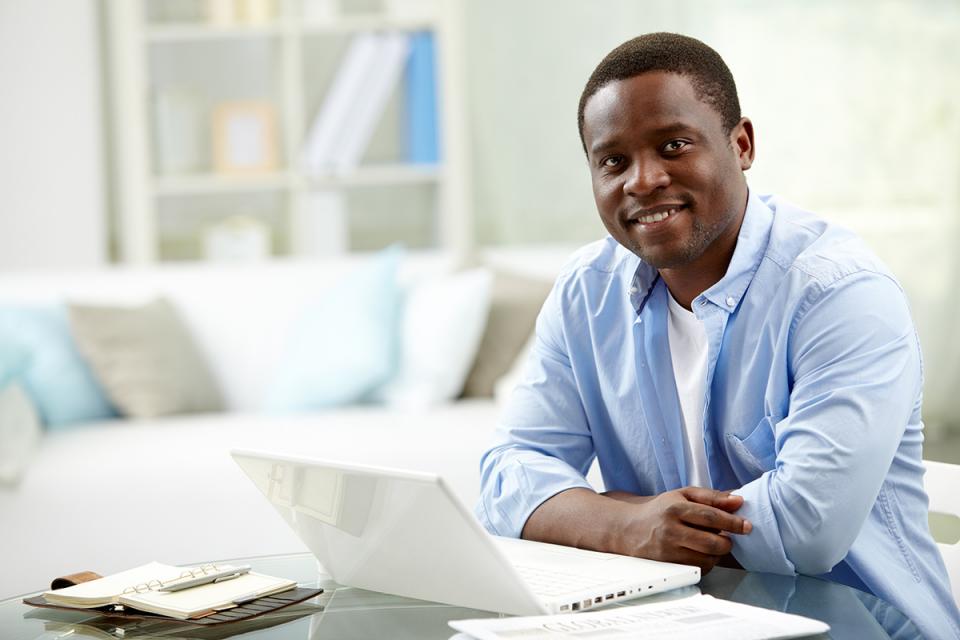 A black man with a smile sits at a table, focused on his laptop.