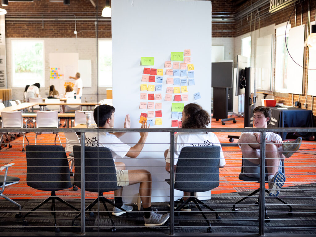 Three students work to refine an idea using sticky notes and a wall