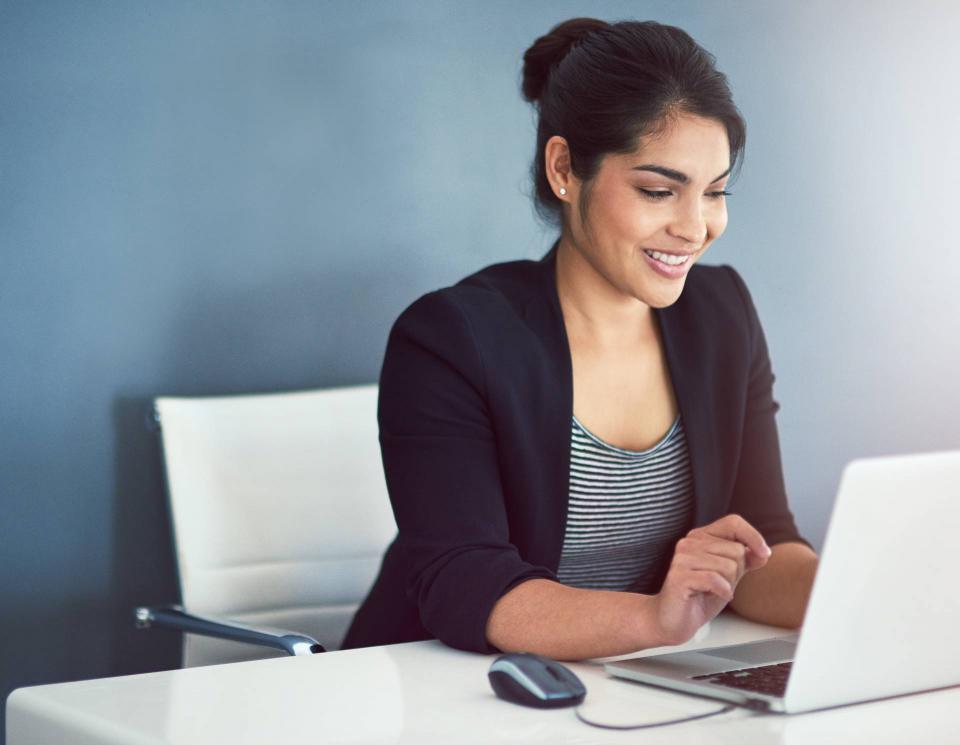 A woman wearing a blue jacket smiles while looking at a laptop at her desk.