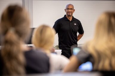 A man in a black shirt standing in front of a classroom, ready to deliver a lecture.