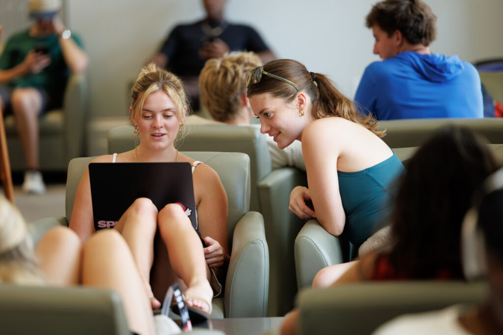 University of Georgia fall semester begins Two individuals sit closely together in a lounge, one using a laptop while the other leans in to observe, surrounded by other people in the background. University of Georgia fall semester begins