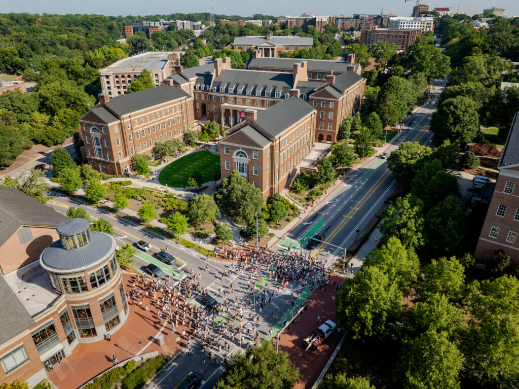 An aerial view shows a large crowd gathered on a street in front of brick university buildings surrounded by lush green trees. University of Georgia fall semester begins