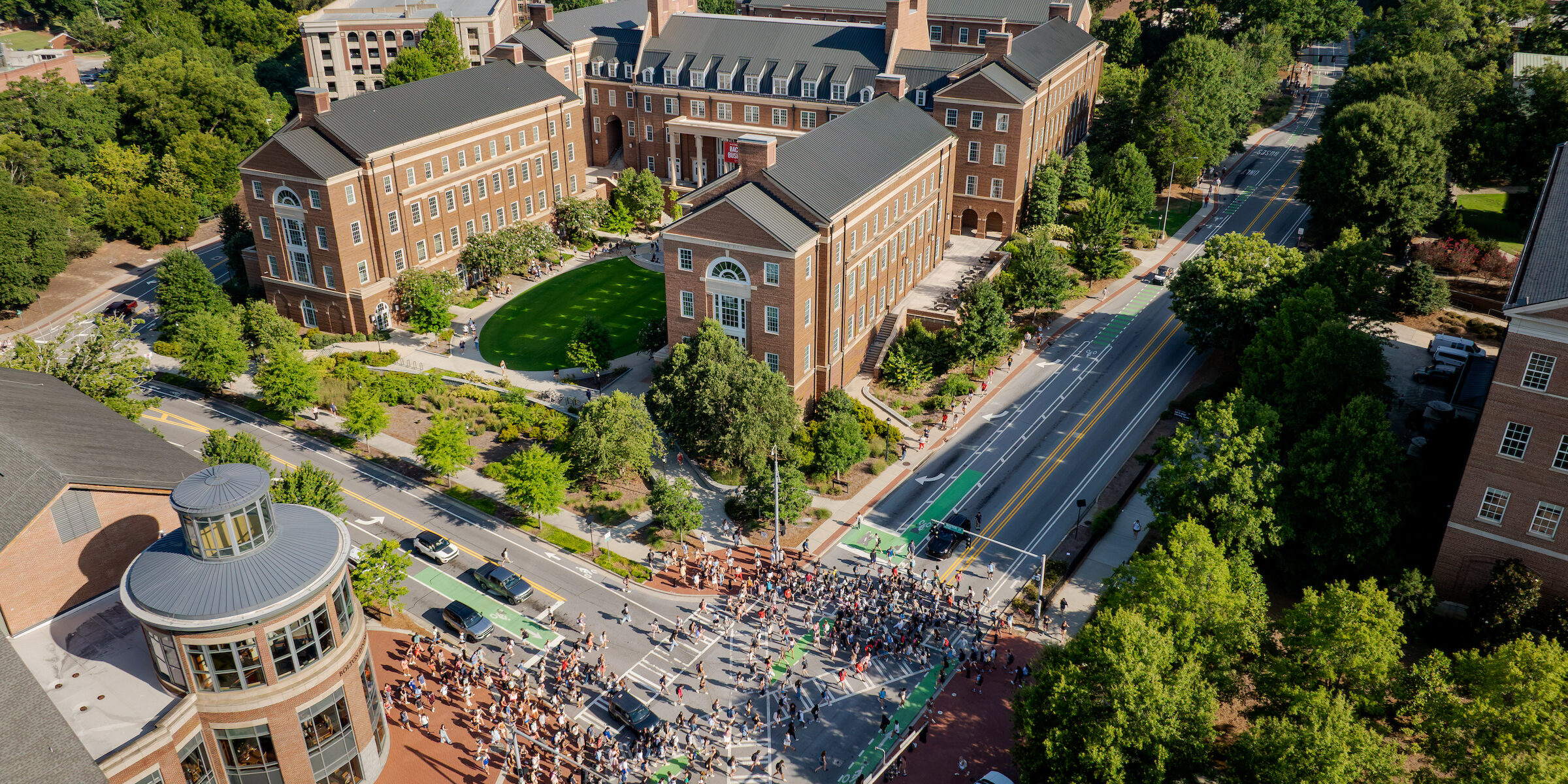 An aerial view shows a large crowd gathered on a street in front of brick university buildings surrounded by lush green trees.