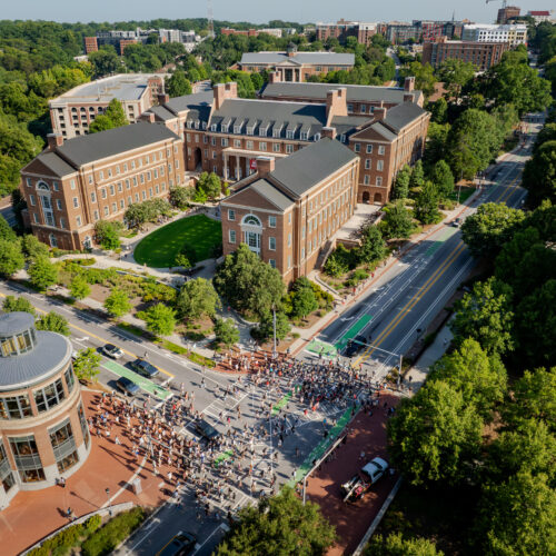 An aerial view shows a large crowd gathered on a street in front of brick university buildings surrounded by lush green trees. University of Georgia fall semester begins