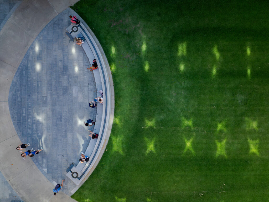 First Day Gallery 6 An overhead view shows people relaxing on a curved stone bench beside a vibrant green lawn, with shadows cast across the grass.