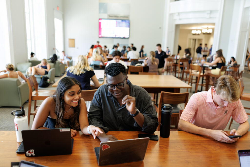First Gallery9 Three students collaborate at a wooden table in a bright, bustling library or study hall, surrounded by other learners and large windows.