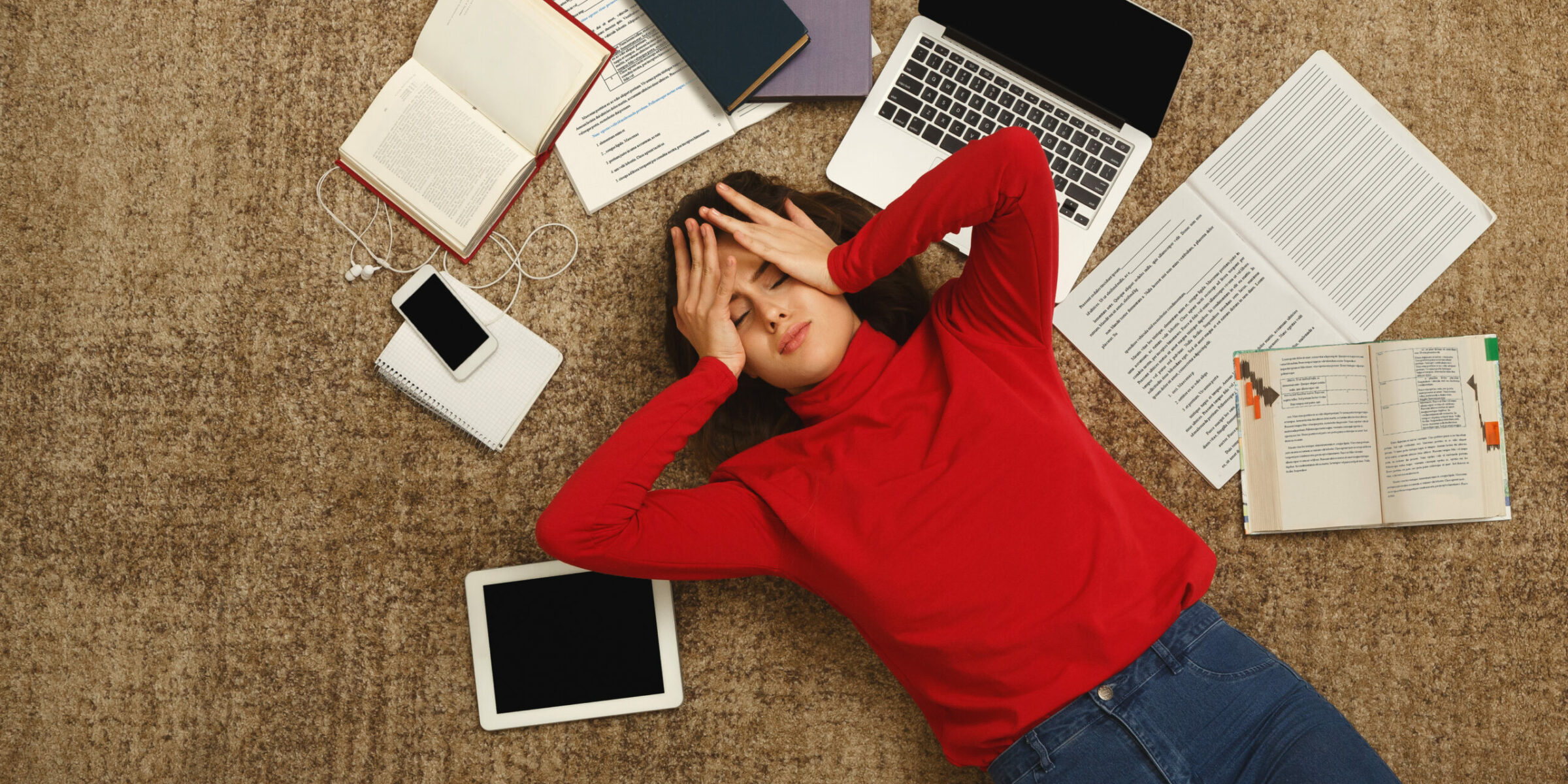 Exhausted student girl lying on the floor among textbooks, tests and gadgets, copy space. Woman holding head with hands.