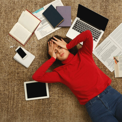 Exhausted student girl lying on the floor among textbooks, tests and gadgets, copy space. Woman holding head with hands.