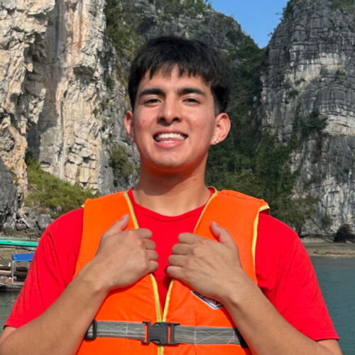 Miguel Martinez in Ha Long Bay Vietnam wearing a red shirt and orange lifevest with mountains and water in the background.