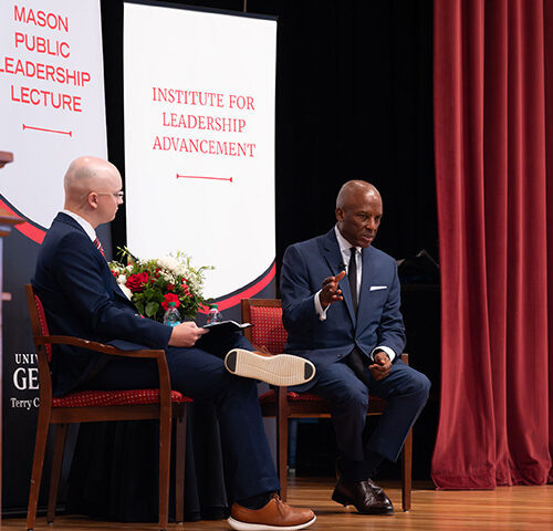 Two individuals in suits are seated on a stage during a public lecture event, with banners displaying "Mason Public Leadership Lecture" and "Institute for Leadership Advancement" behind them.