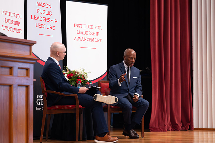 Two individuals in suits are seated on a stage during a public lecture event, with banners displaying "Mason Public Leadership Lecture" and "Institute for Leadership Advancement" behind them.