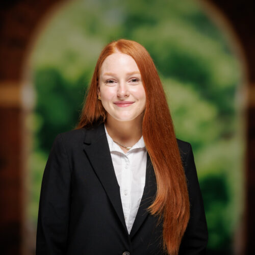 A person with long red hair stands in a black blazer and white shirt, smiling softly in front of an arched doorway with a blurred green background.