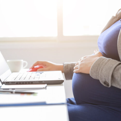 A woman in a blazer places her hand on her pregnant belly while working on her laptop