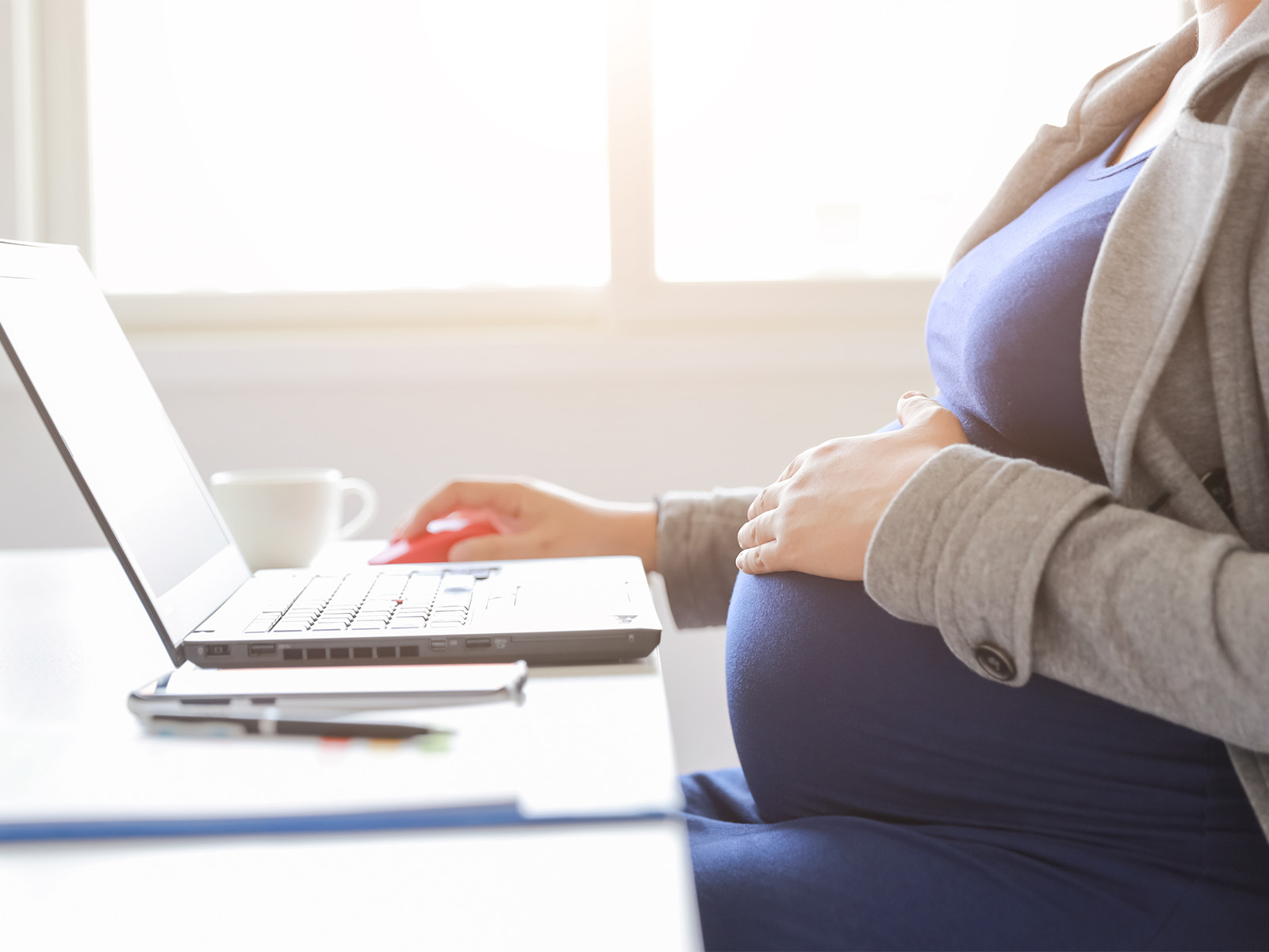 A woman in a blazer places her hand on her pregnant belly while working on her laptop