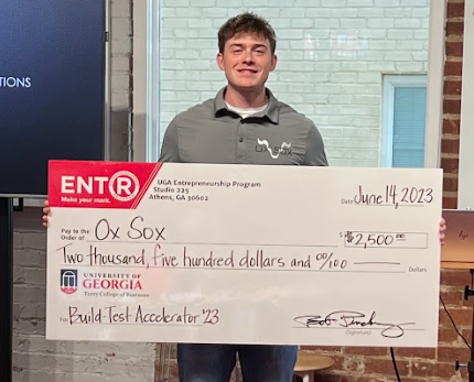 A male student in a gray polo shirt holds a large check for $2,500 after his company, Ox Sox, won the Build Test Accelerator competition hosted by the Terry College of Business.