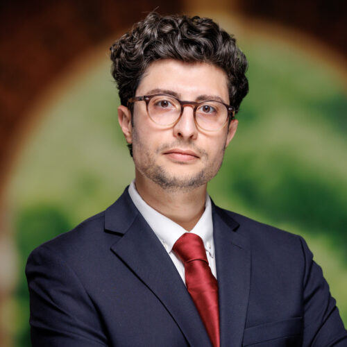 A person with curly hair and glasses, wearing a dark suit and red tie, stands with arms crossed in front of an arched doorway.