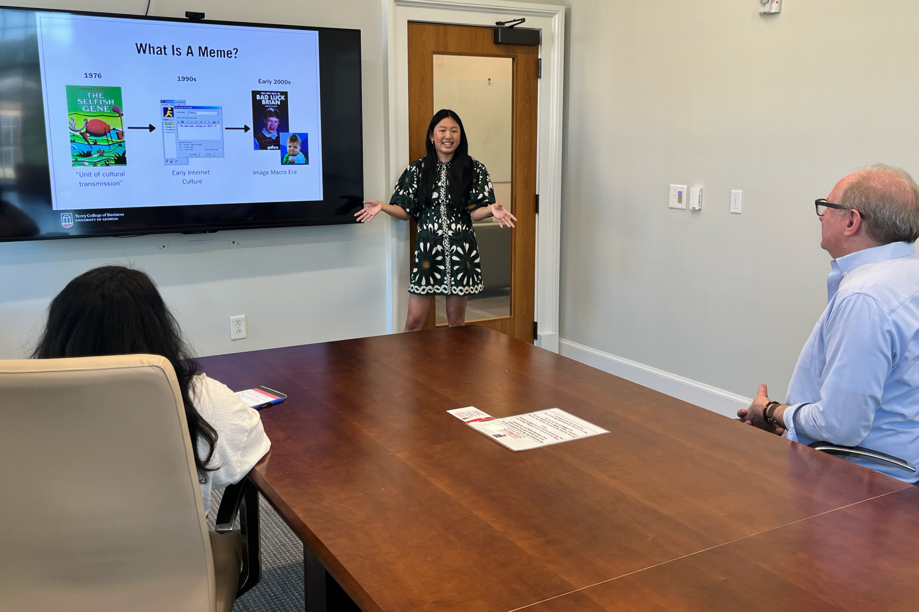 Terry College Undergraduate Researcher Sophia Beasley stands in front of large conference room's screen with a Power Point slide that reads "What is a meme?",