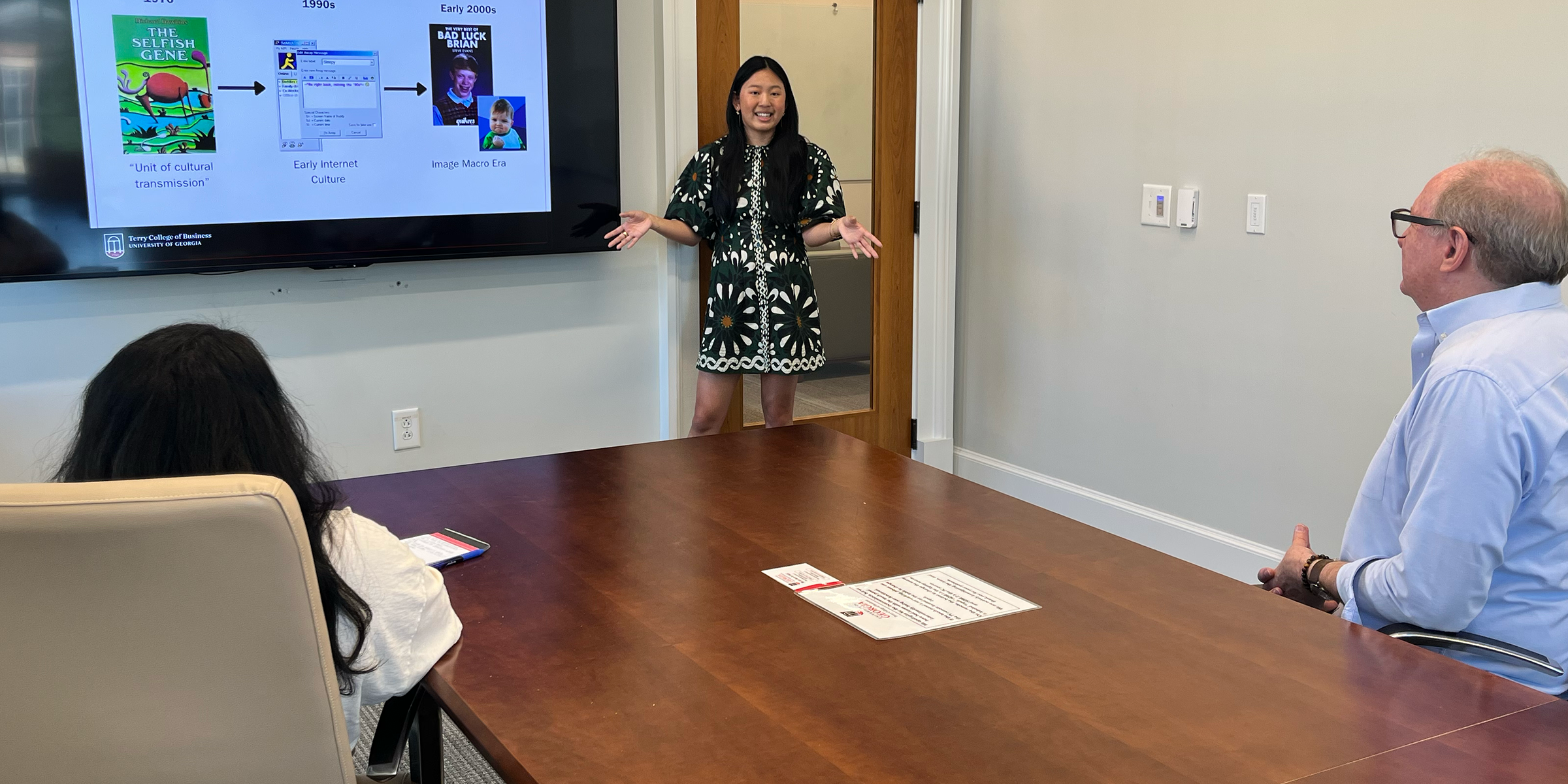 Terry College Undergraduate Researcher Sophia Beasley stands in front of large conference room's screen with a Power Point slide that reads "What is a meme?",